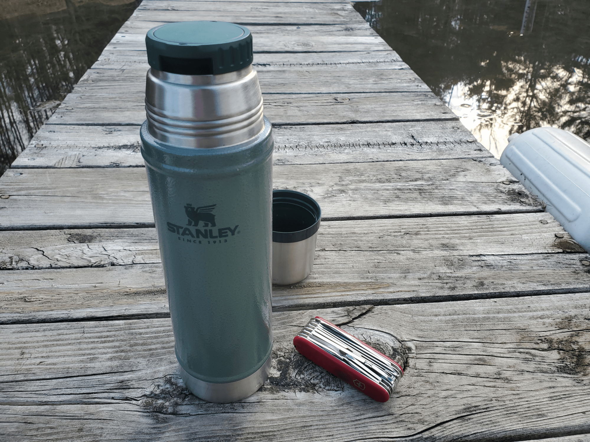 Photo of a Stanley Thermos and a Victorinox Swiss Champ knife on a dock along a lake.